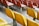 A view of several rows of empty stadium seating, featuring bright yellow plastic chairs in the foreground and red ones in the background. The seating is mounted on metal poles and the concrete flooring is visible beneath.