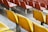 A view of several rows of empty stadium seating, featuring bright yellow plastic chairs in the foreground and red ones in the background. The seating is mounted on metal poles and the concrete flooring is visible beneath.