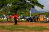 A bustling outdoor scene with people gathered around a parked yellow auto rickshaw. A man in a red shirt walks toward the forefront, carrying objects in his arms. Several people are standing near bicycles and cars, with a backdrop of lush green trees and a distant mountain under a clear sky.