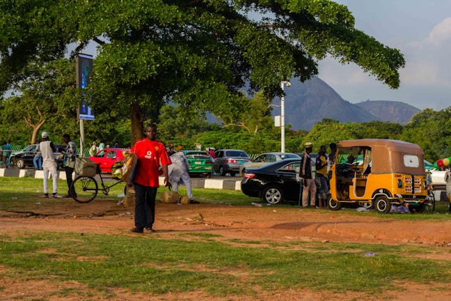 A bustling outdoor scene with people gathered around a parked yellow auto rickshaw. A man in a red shirt walks toward the forefront, carrying objects in his arms. Several people are standing near bicycles and cars, with a backdrop of lush green trees and a distant mountain under a clear sky.