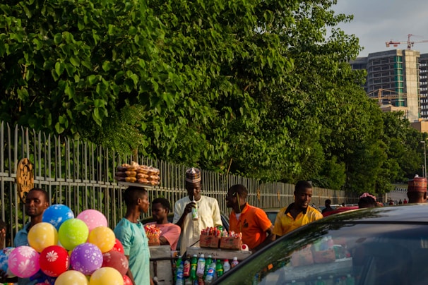 A bustling street scene with several people engaged in selling goods. The area is lively, featuring street vendors carrying colorful balloons, trays of snacks, and bottles of beverages. Green foliage is abundant, lining a metal fence, and a tall building under construction is visible in the background.