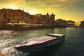 A sleek speedboat with red and white stripes floats on calm waters near a coastal town. The background features a picturesque view of colorful buildings with distinctive architecture, a church with a dome, and a setting sun casting a golden hue over the scene. The water reflects the warm light, adding a shimmering effect.