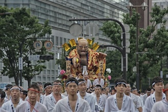 A group of people dressed in traditional white garments are participating in a festival parade, carrying a large, elaborate float. The float features a large, vibrant figure with a fierce expression, adorned with golden and red elements. The scene takes place in a city with tall buildings and greenery visible in the background.