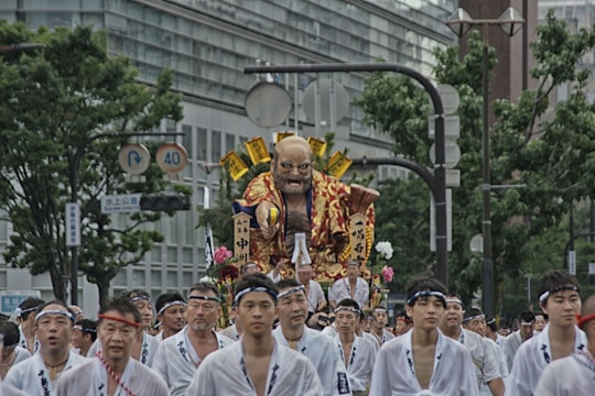 A group of people dressed in traditional white garments are participating in a festival parade, carrying a large, elaborate float. The float features a large, vibrant figure with a fierce expression, adorned with golden and red elements. The scene takes place in a city with tall buildings and greenery visible in the background.