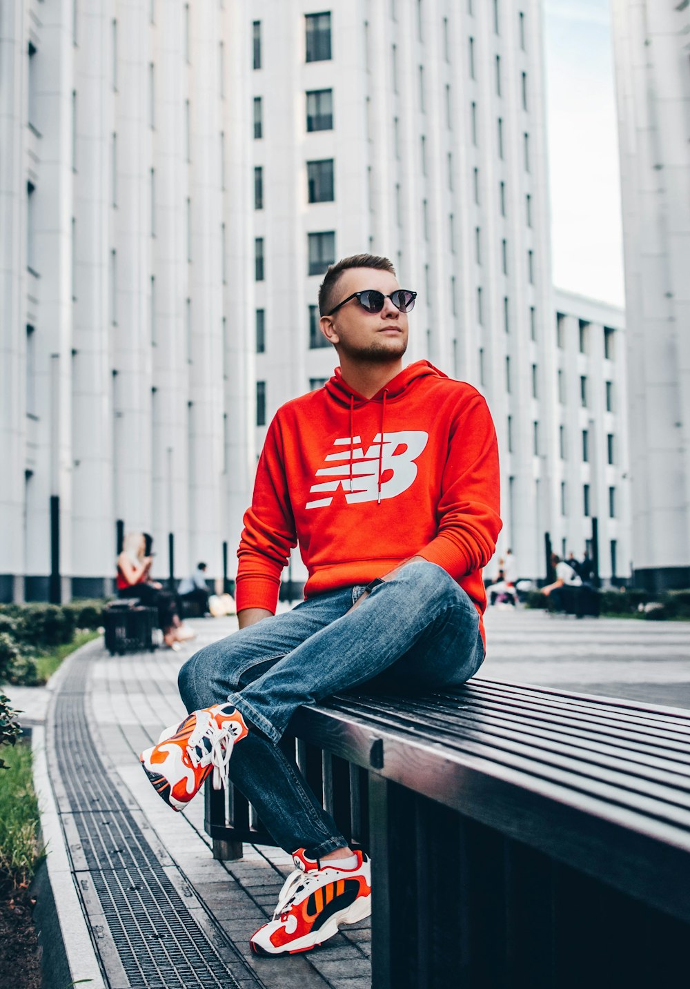 man wearing red New Balance pullover hoodie sitting on black bench in front of white high-rise building