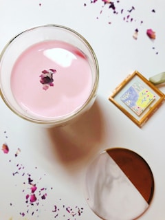 A glass filled with a pink liquid is placed on a surface, with dried flower petals floating on top. Nearby, a small framed artwork with abstract designs sits on the right. Scattered petals are visible around the glass, adding a decorative touch.
