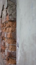 A close-up view of an old brick wall with cracked and peeling plaster. The bricks are exposed as the plaster has fallen away, revealing the texture and color of the red and brown bricks beneath.