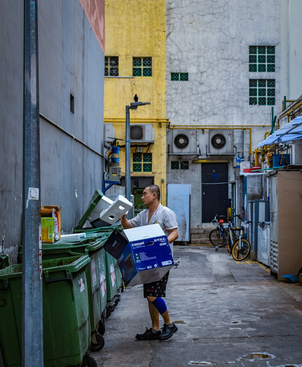 Hombre con camisa gris abotonada sosteniendo una caja azul cerca de un cubo de basura verde