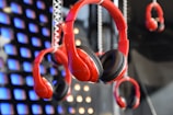 A display of multiple red headphones hanging in front of a backdrop featuring illuminated blue and orange squares. The focus is on the central pair of headphones, which are prominently featured among others in the background.