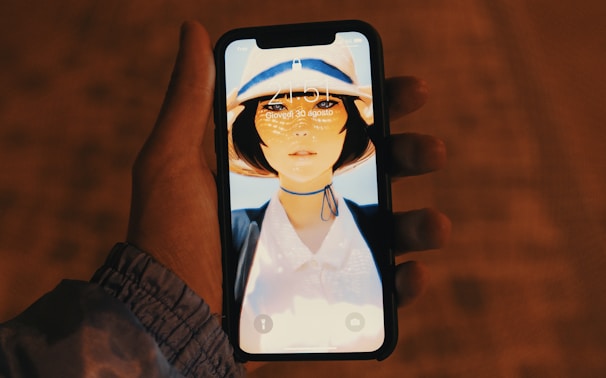 A hand is holding a smartphone showing a lock screen with an image of a woman wearing a hat and a choker. The setting appears to be outdoors during the evening or nighttime, given the warm, low lighting in the background.