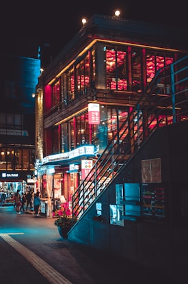 A vibrant urban scene at night featuring a multi-story building with large glass windows illuminated by warm yellow and red lights. The street level displays a variety of bright neon signs in different colors, while a staircase ascends to the upper levels. People are walking along the sidewalk, adding life to the atmosphere. The overall setting suggests a lively and bustling nightlife environment.