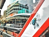 A close-up of a storefront window with various nail polish bottles arranged in rows behind the glass. The reflection of a street scene is visible, showing a modern building and a person walking. A large illustration of a fly is prominently displayed on the window.