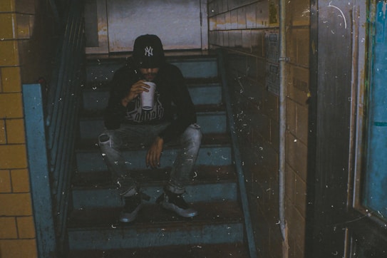 A person wearing a dark hoodie, jeans, and a baseball cap is sitting on dimly lit stairs, drinking from a cup. The setting appears to be an indoor stairway, with blue and yellow tiled walls and an overall gritty, urban atmosphere.