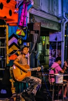 A street performer is playing an acoustic guitar while seated under vibrant, colorful decorations. The performer is in front of a storefront with signs in both English and Arabic. Another person is seated nearby, clapping along. A tip bucket is visible on a stool beside the performer.