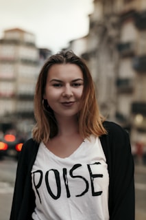 A woman with shoulder-length brown hair is wearing a white t-shirt with the word 'POISE' printed on it. She stands outdoors in an urban setting with blurred buildings and traffic lights in the background, giving the scene a city vibe.