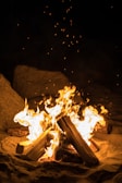 Logs arranged in a pyramid shape burn brightly in the center, with flames reaching upward and sparks flying. The fire sits on a sandy surface surrounded by large rocks, creating a natural outdoor setting.