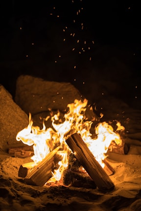 Logs arranged in a pyramid shape burn brightly in the center, with flames reaching upward and sparks flying. The fire sits on a sandy surface surrounded by large rocks, creating a natural outdoor setting.