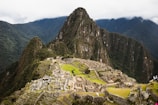 Ruins of an ancient Incan city set against a backdrop of towering green mountains and a cloudy sky. The structures are made of stone and are arranged in a terraced fashion, with some parts showing greenery.