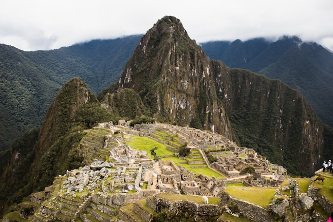 Ruins of an ancient Incan city set against a backdrop of towering green mountains and a cloudy sky. The structures are made of stone and are arranged in a terraced fashion, with some parts showing greenery.