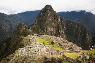 Ruins of an ancient Incan city set against a backdrop of towering green mountains and a cloudy sky. The structures are made of stone and are arranged in a terraced fashion, with some parts showing greenery.