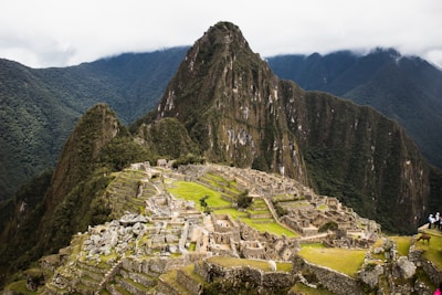 Ruins of an ancient Incan city set against a backdrop of towering green mountains and a cloudy sky. The structures are made of stone and are arranged in a terraced fashion, with some parts showing greenery.