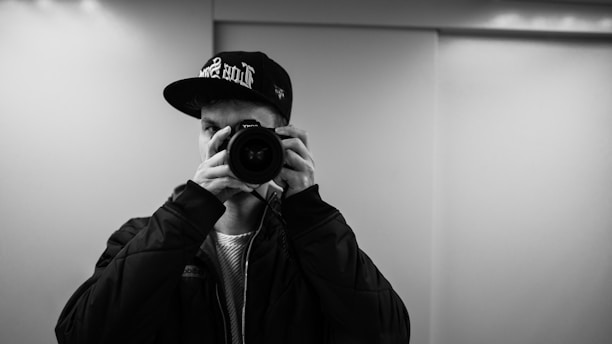 A person wearing a baseball cap and a jacket holds a camera up to their face, capturing a photo. The setting appears to be indoors with soft lighting and a blank wall in the background.