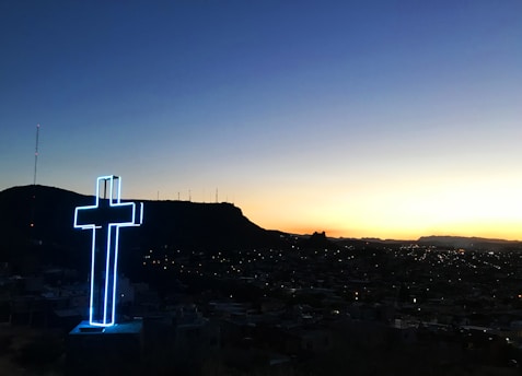 A large, illuminated cross stands prominently overlooking a cityscape during sunset. The sky transitions smoothly from deep blue to a warm golden hue near the horizon. Silhouettes of distant hills and scattered city lights offer a tranquil and serene setting.