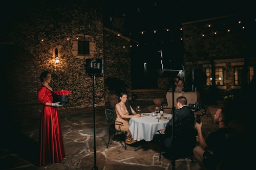 An outdoor nighttime scene with a romantic ambiance captured in a courtyard. A woman in a striking red dress holds a bouquet of red roses, standing near a couple dining at a small, round table under soft lighting. The setting includes stone walls and hanging string lights, and a crew member is filming the couple, suggesting a photoshoot or a film scene.