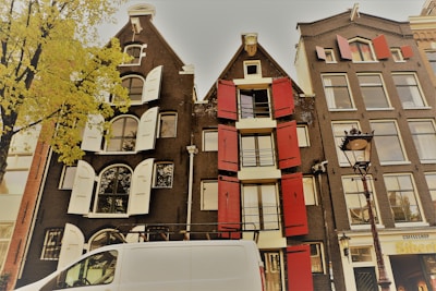 A street view featuring traditional European buildings with distinct architectural features such as red and white shutters. The buildings are multi-story with large windows. A white van is parked in front, and a decorative street lamp is visible. A tree with yellow-green leaves is on the left, and a small shop sign is partially visible on the right.