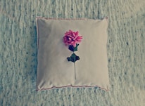 A square cushion with a plush texture lies on a soft, textured surface. Centered on the cushion is a single, vibrant pink flower with a green stem and leaves.