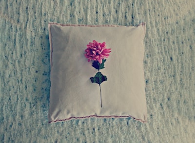 A square cushion with a plush texture lies on a soft, textured surface. Centered on the cushion is a single, vibrant pink flower with a green stem and leaves.