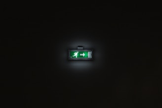 A glowing green emergency exit sign is centered within a dark background. The sign features a person running and an arrow pointing to the right.