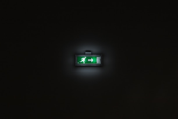 A glowing green emergency exit sign is centered within a dark background. The sign features a person running and an arrow pointing to the right.