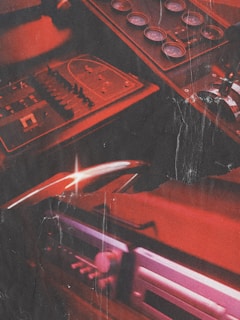 A dimly lit control panel with numerous dials, gauges, and switches. The overall color scheme features a strong red hue, giving the scene an intense, industrial appearance. The panel appears to be part of a vintage or retro setup, and the lighting creates reflective highlights across the surfaces.