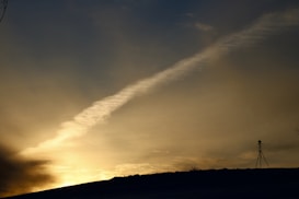 A vast expanse of sky is painted with warm hues of sunset, creating a gradient from deep orange to soft yellow. A striking contrail stretches diagonally across the sky. Silhouetted against this backdrop, a lone camera tripod stands on a hill, adding a sense of solitude and focus.