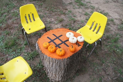 A large tree stump has been converted into a tic-tac-toe game with black grid lines painted on it. Miniature pumpkins, both orange and white, are placed on the grid as game pieces. Three yellow plastic chairs are positioned around the stump on a grassy and dirt-covered outdoor surface.