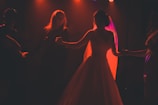 A silhouette of a bride in a flowing gown dancing with a group of people. The scene is dramatically lit with warm, orange-red lights casting an ethereal glow around the bride. The background is dark, highlighting the figures and creating a sense of intimacy.