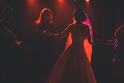 A silhouette of a bride in a flowing gown dancing with a group of people. The scene is dramatically lit with warm, orange-red lights casting an ethereal glow around the bride. The background is dark, highlighting the figures and creating a sense of intimacy.