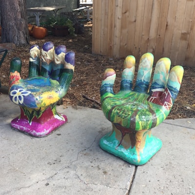 Two large, hand-shaped sculptures are positioned on an outdoor surface, painted with vibrant colors depicting natural scenes. The left sculpture features floral designs and mountainous landscapes, while the right one includes carvings of a barn and fields. The setting includes pumpkins and wooden structures, indicating a rustic, outdoor environment.