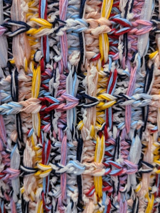 An intricate pattern of multicolored yarns intricately woven together, forming a textured surface. The yarns are intertwined in a complex arrangement, showcasing a variety of colors including pastel pinks, blues, and yellows.