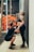 A man kneels in front of a woman, holding her hips, as she leans back against a wall, laughing. Both individuals are dressed in athletic wear, and the background suggests a gym environment with gym equipment and ropes.