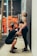 A man kneels in front of a woman, holding her hips, as she leans back against a wall, laughing. Both individuals are dressed in athletic wear, and the background suggests a gym environment with gym equipment and ropes.