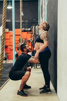 A man kneels in front of a woman, holding her hips, as she leans back against a wall, laughing. Both individuals are dressed in athletic wear, and the background suggests a gym environment with gym equipment and ropes.