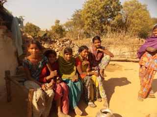 A group of young people are gathered, seated casually on a wooden bench or structure outdoors in a rustic setting. They are wearing colorful traditional clothing, and the scene is bathed in warm sunlight. Behind them, there is a backdrop of trees and what appears to be a fence made of wooden sticks, as well as stacks of stones or firewood.