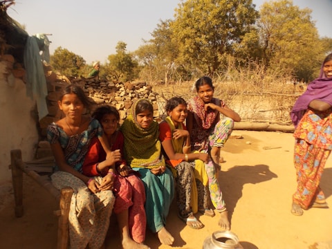 A group of young people are gathered, seated casually on a wooden bench or structure outdoors in a rustic setting. They are wearing colorful traditional clothing, and the scene is bathed in warm sunlight. Behind them, there is a backdrop of trees and what appears to be a fence made of wooden sticks, as well as stacks of stones or firewood.