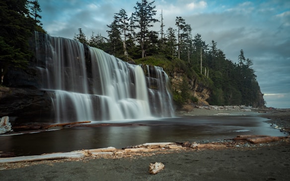 A serene waterfall cascades gently down a rocky cliff, surrounded by lush evergreen trees. The water flows into a calm pool, reflecting the surrounding landscape. Driftwood is scattered along the sandy shore, adding a natural element to the scene. The sky is overcast, hinting at a peaceful yet moody atmosphere.
