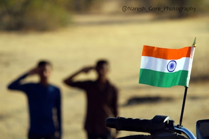 Two blurred individuals in the background are saluting, with an Indian flag in sharp focus in the foreground. The flag, attached to a motorcycle handlebar, waves gently.