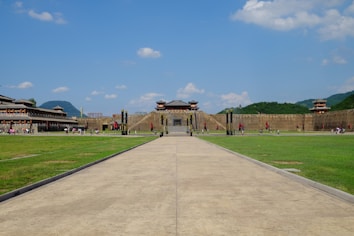 An expansive view of a historical Chinese complex with traditional architecture, featuring tall walls, structures with tiered roofs, and red flags lining a long path. The area is situated amidst green hills under a clear blue sky with scattered clouds, and people can be seen walking and enjoying the space.