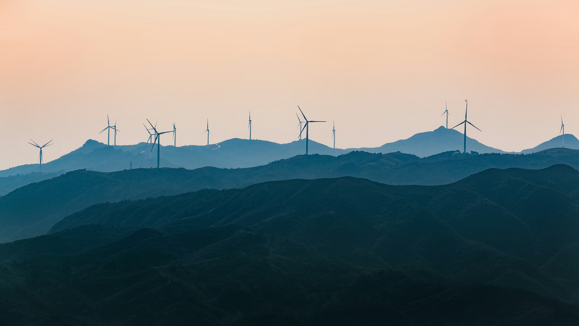 A row of distant hills with a light blue haze over them, studded with wind farms, before a pink-orange sky