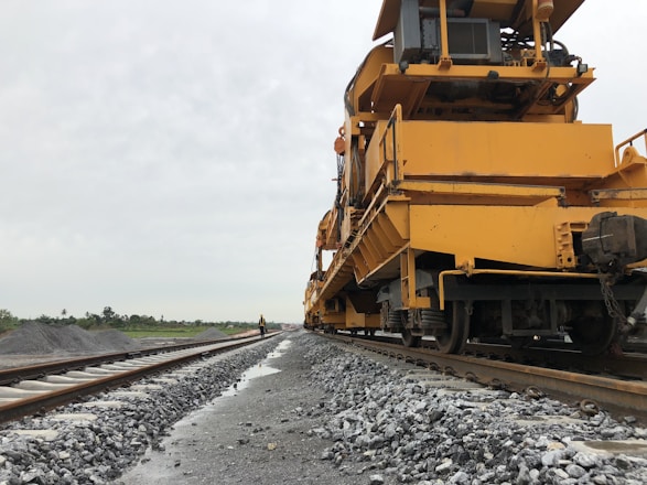 A large, yellow maintenance machine is positioned on the railway tracks, extending along the length of the tracks. The backdrop features some greenery under an overcast sky, and a person in an orange vest stands further down the tracks.